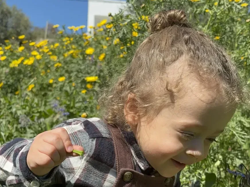 Un petit enfant aux cheveux bouclés sourit en cueillant une fleur jaune dans un champ.